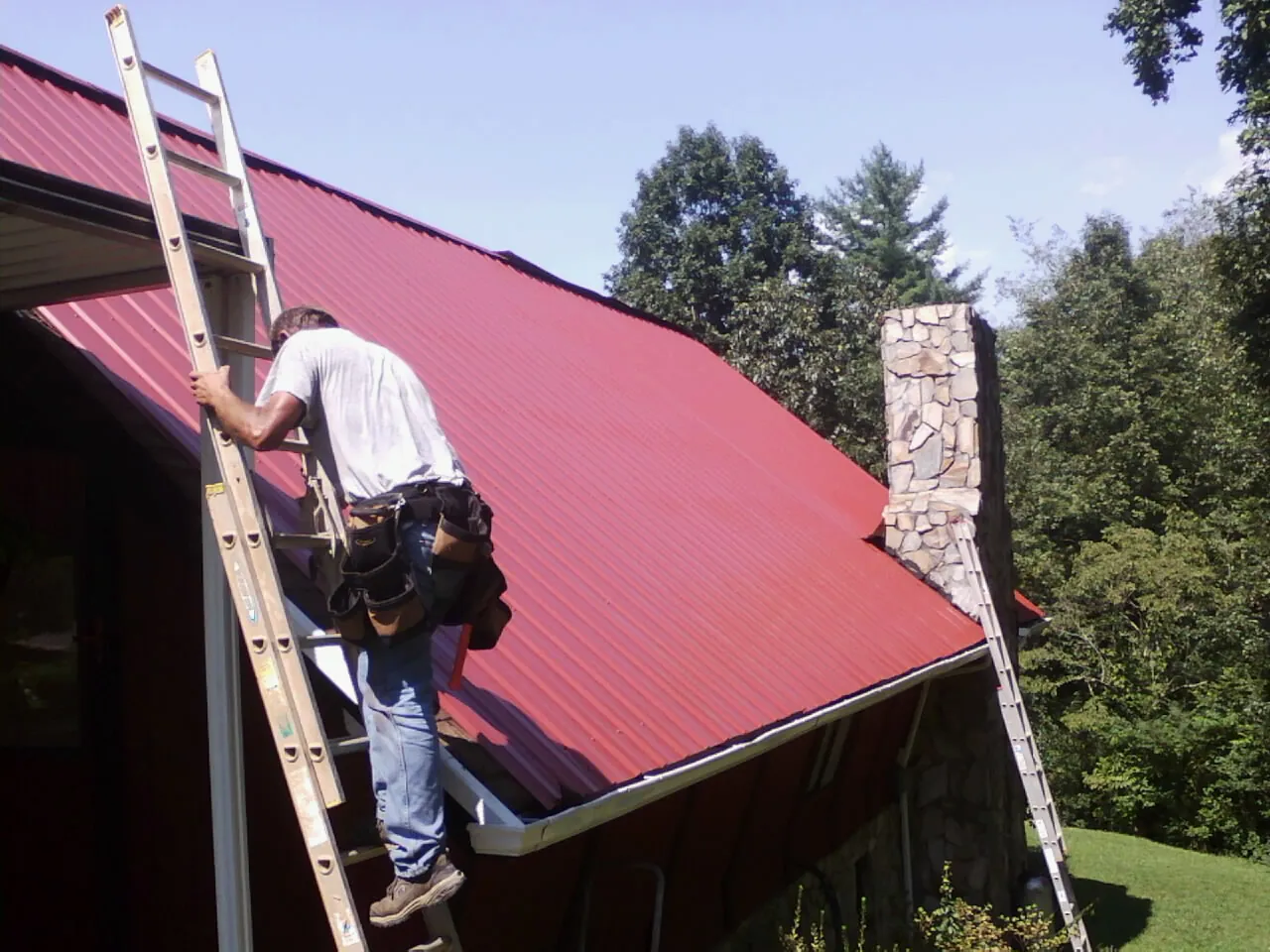 Roofer on a ladder beside a red metal roof and stone chimney — Soffit & Fascia Repair by Roofing Brothers in Elton